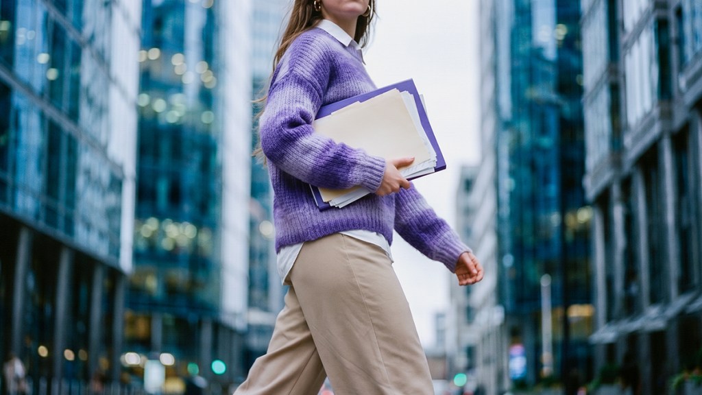 Mujer profesional con suéter morado sosteniendo una carpeta de documentos en una calle urbana rodeada de edificios corporativos.