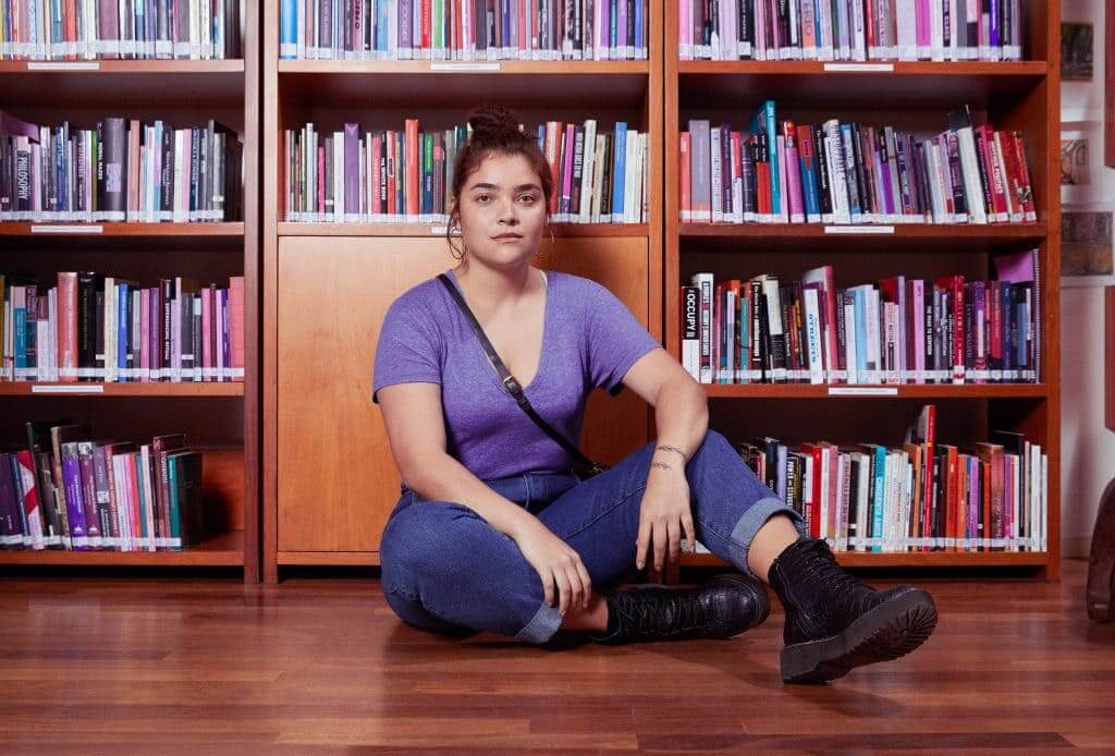 Mujer pelirroja de mediana edad usa una playera morada y jeans con botas negras, se encuentra sentada en el piso de una biblioteca meditando acerca de dinero y cuestiones de finanzas personales para la mujer