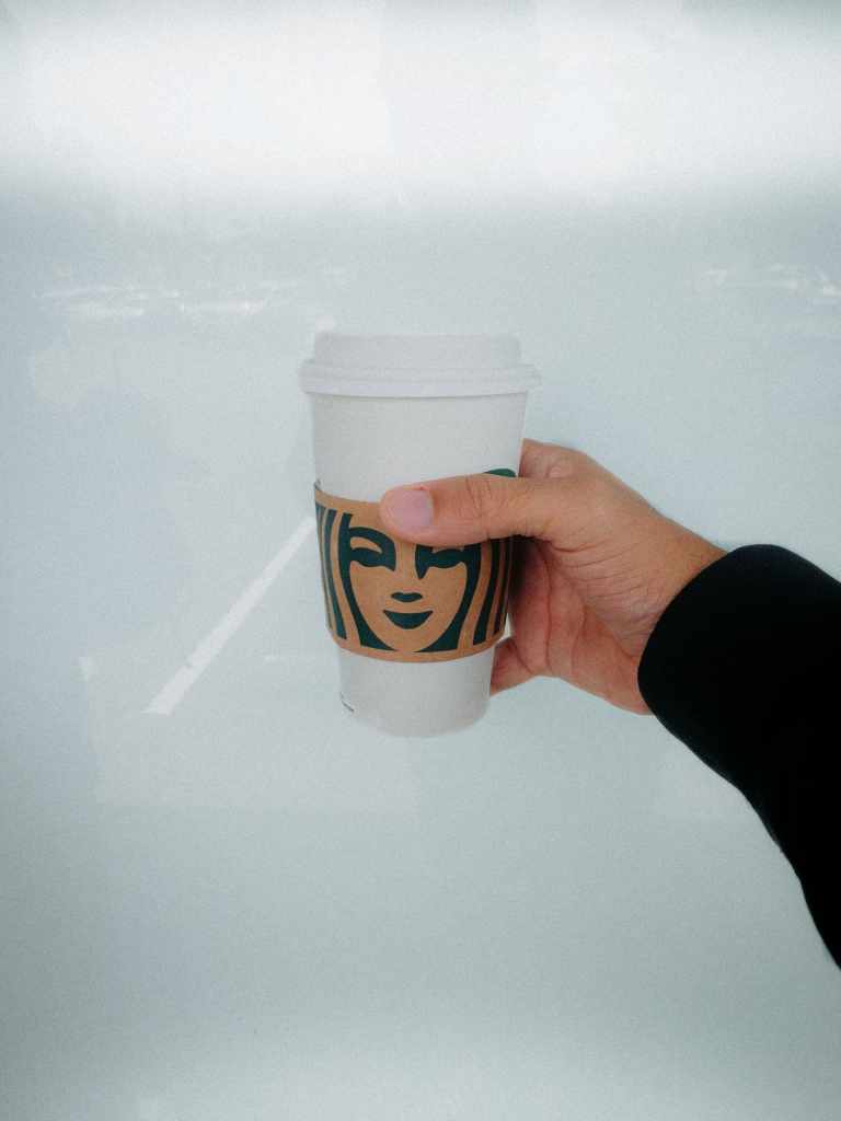 Mano sostiene un café en vaso desechable frente a una ventana ejemplificando uno de los hábitos que no te dejan ahorrar dinero