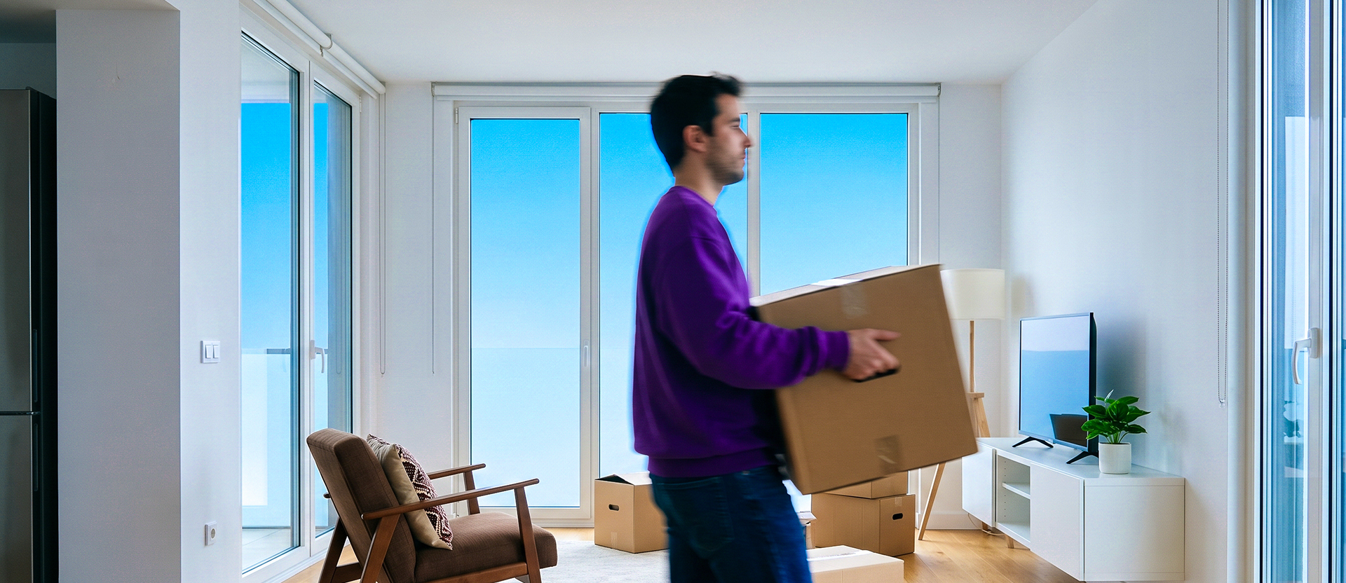 Persona con suéter morado cargando una caja de cartón durante una mudanza en un apartamento moderno con vista al mar.