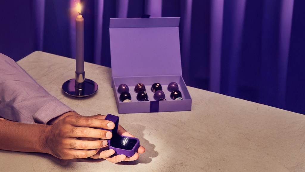 las manos de un joven abren una caja de terciopelo morado con un anillo de compromiso sobre una mesa color café junto a una vela morada encendida y una caja chocolates en un ambiente acogedor que invita a cuestionar cuánto cuesta casarse en méxico