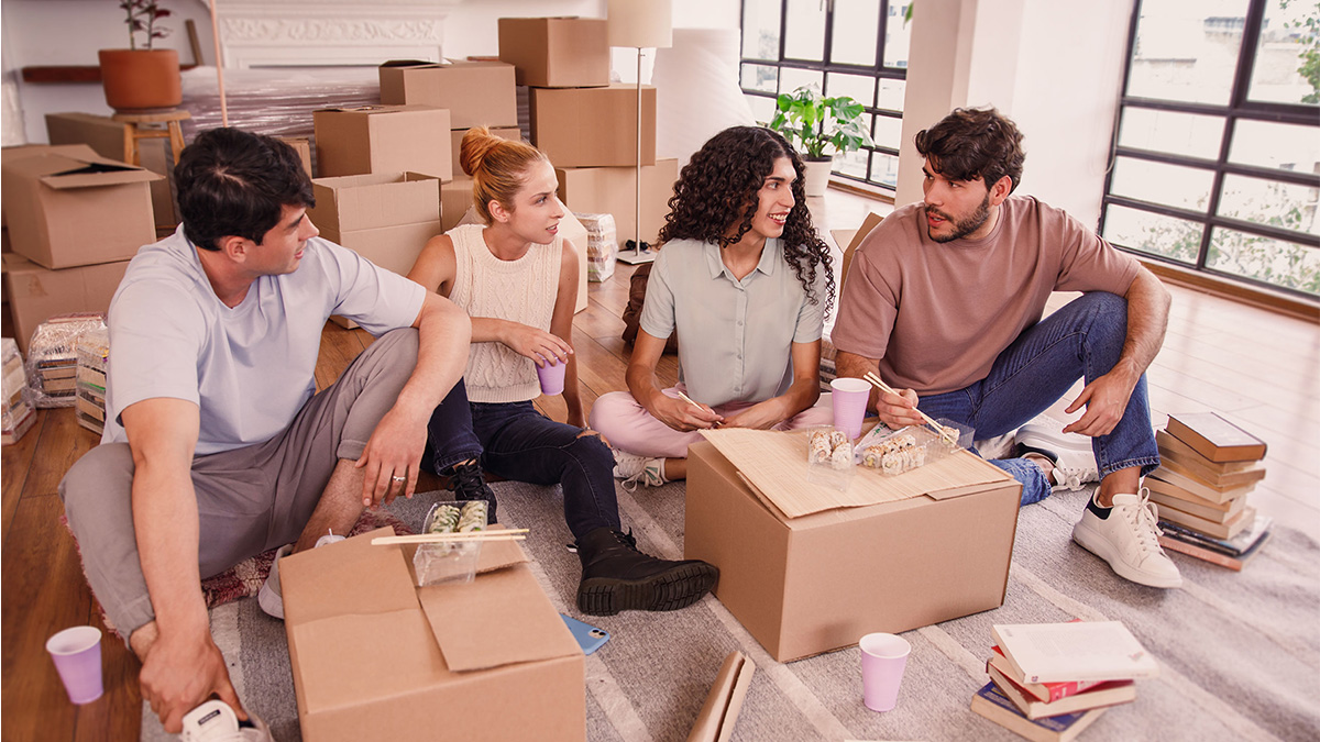 Cuatro chicos de aproximadamente 25 años vestidos con ropa casual toman un café sobre cajas de cartón que sugieren una mudanza mientras hablan sobre ahorro colectivo