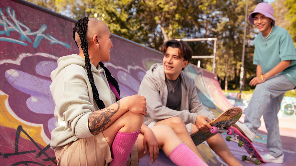 Dos jóvenes en sus veintes vestidos con ropa gris y rosa de veranos platican sobre la independenci financiera en una rampa de patinaje al interior de un parque público