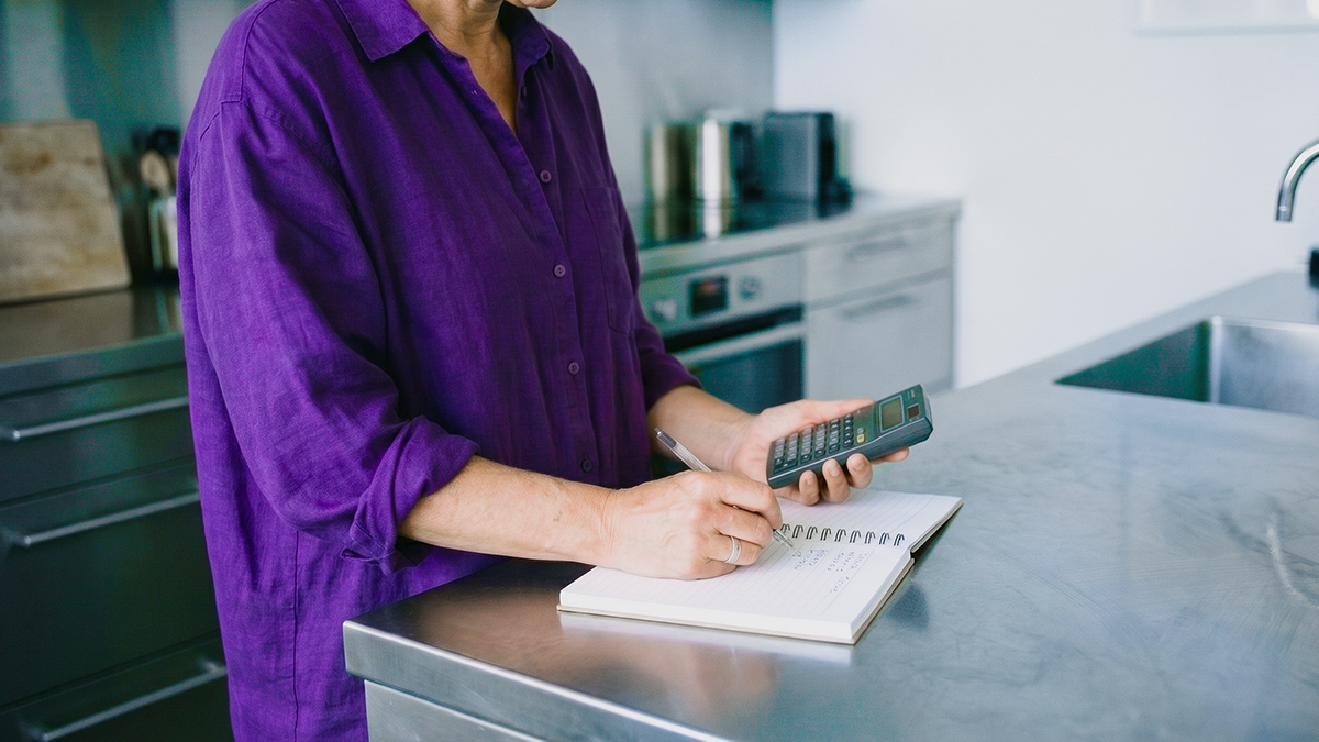 Persona usando una calculadora y anotando en un cuaderno en la cocina, ilustrando cómo funciona el interés compuesto: calcular cómo tus rendimientos se reinvierten y generan nuevos rendimientos automáticamente.