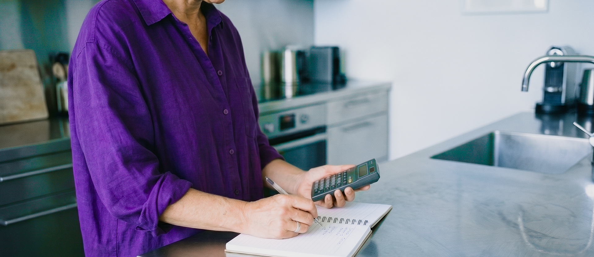 Persona usando una calculadora y anotando en un cuaderno en la cocina, ilustrando cómo funciona el interés compuesto: calcular cómo tus rendimientos se reinvierten y generan nuevos rendimientos automáticamente.