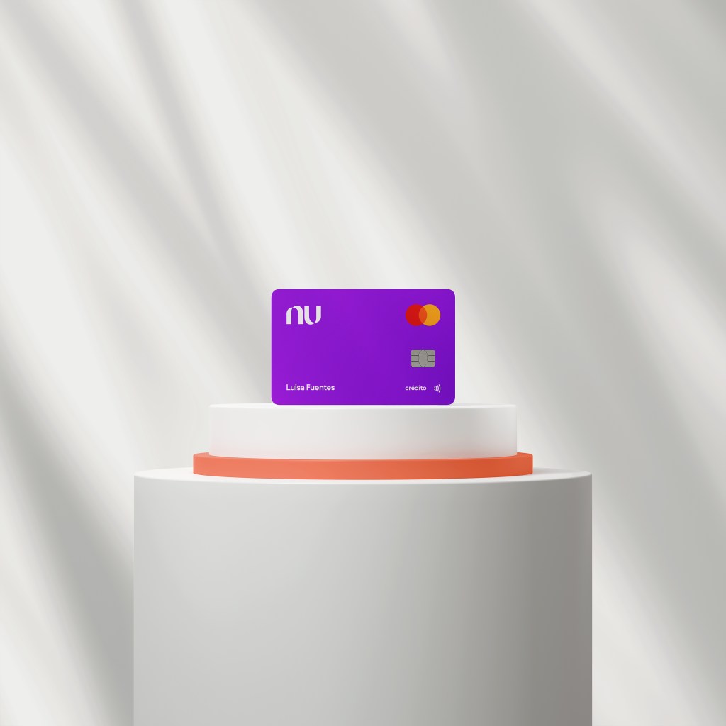 La tarjeta de crédito nu color morado reposa sobre una base de exhibición de color blanco y anaranjado frente a una fondo blanco con sombras suaves. Una tarjeta de crédito sin anualidad, sin comisiones sorpresa ni tarifas abusivas