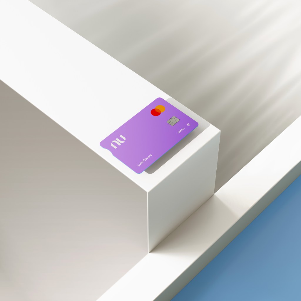 La Tarjeta Cuenta Nu, una cuenta de ahorro que te da altos rendimientos y tarjeta de débito reposa en una base angulada de color blanco frente a un fondo de color azul 