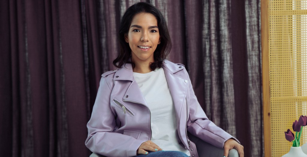 Ximena Salgado, Senior Product Manager viste una chaqueta color morado y una playera blanca mientras está sentada frente a una cortina violeta para explicar como Nu desarrolla sus productos