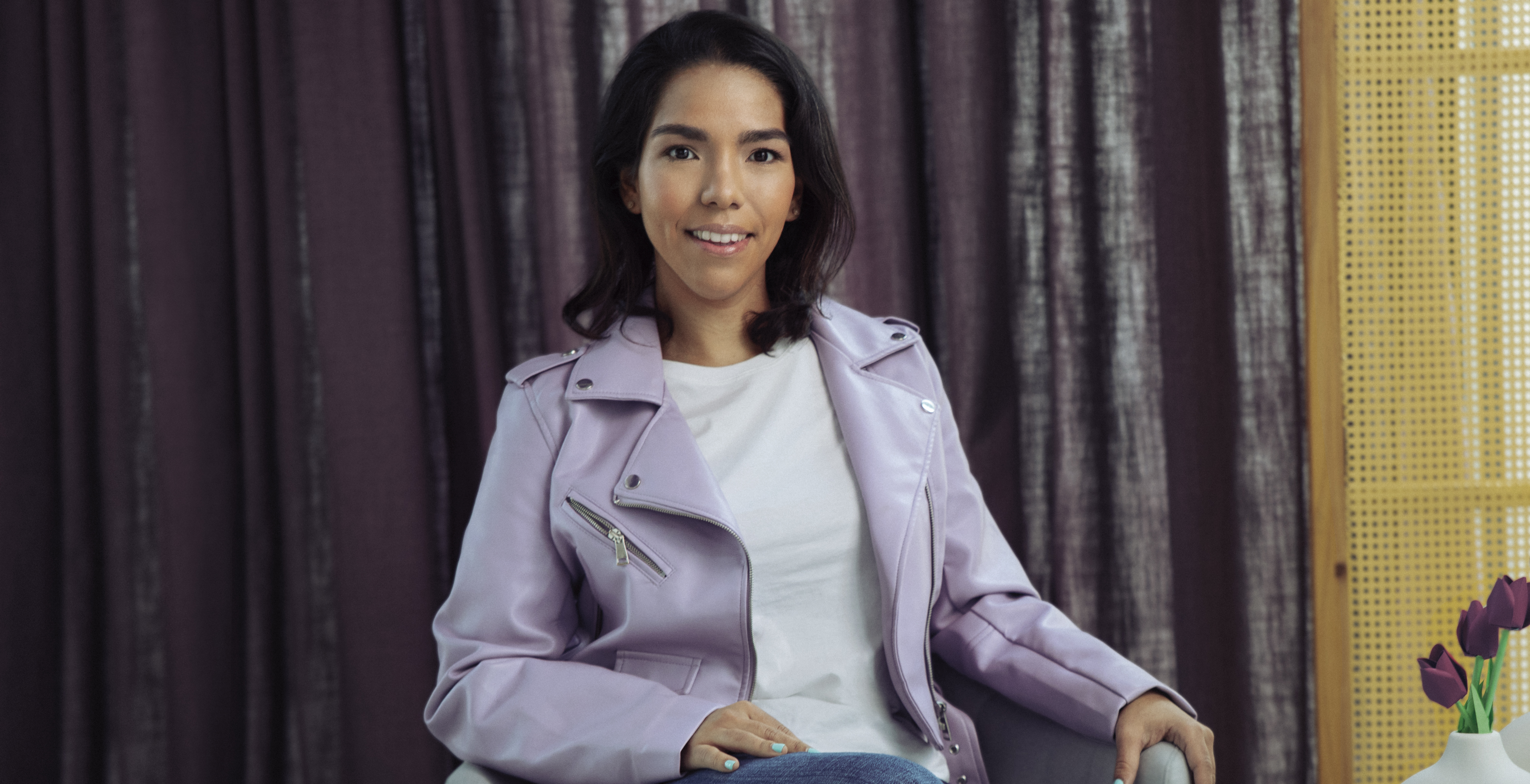 Ximena Salgado, Senior Product Manager viste una chaqueta color morado y una playera blanca mientras está sentada frente a una cortina violeta para explicar como Nu desarrolla sus productos