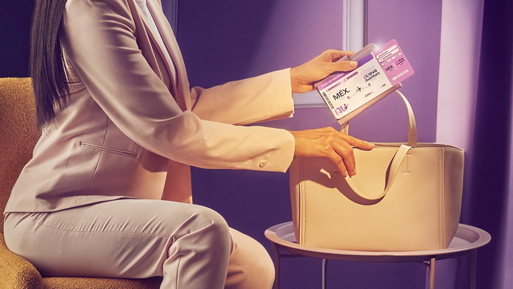 Una mujer con un traje sastre color beige se encuentra sentada en una sala de espera color morado guardando su pase de abordar en un bolso de piel color beige mientras piensa en las mejores tarjetas de crédito para viajar
