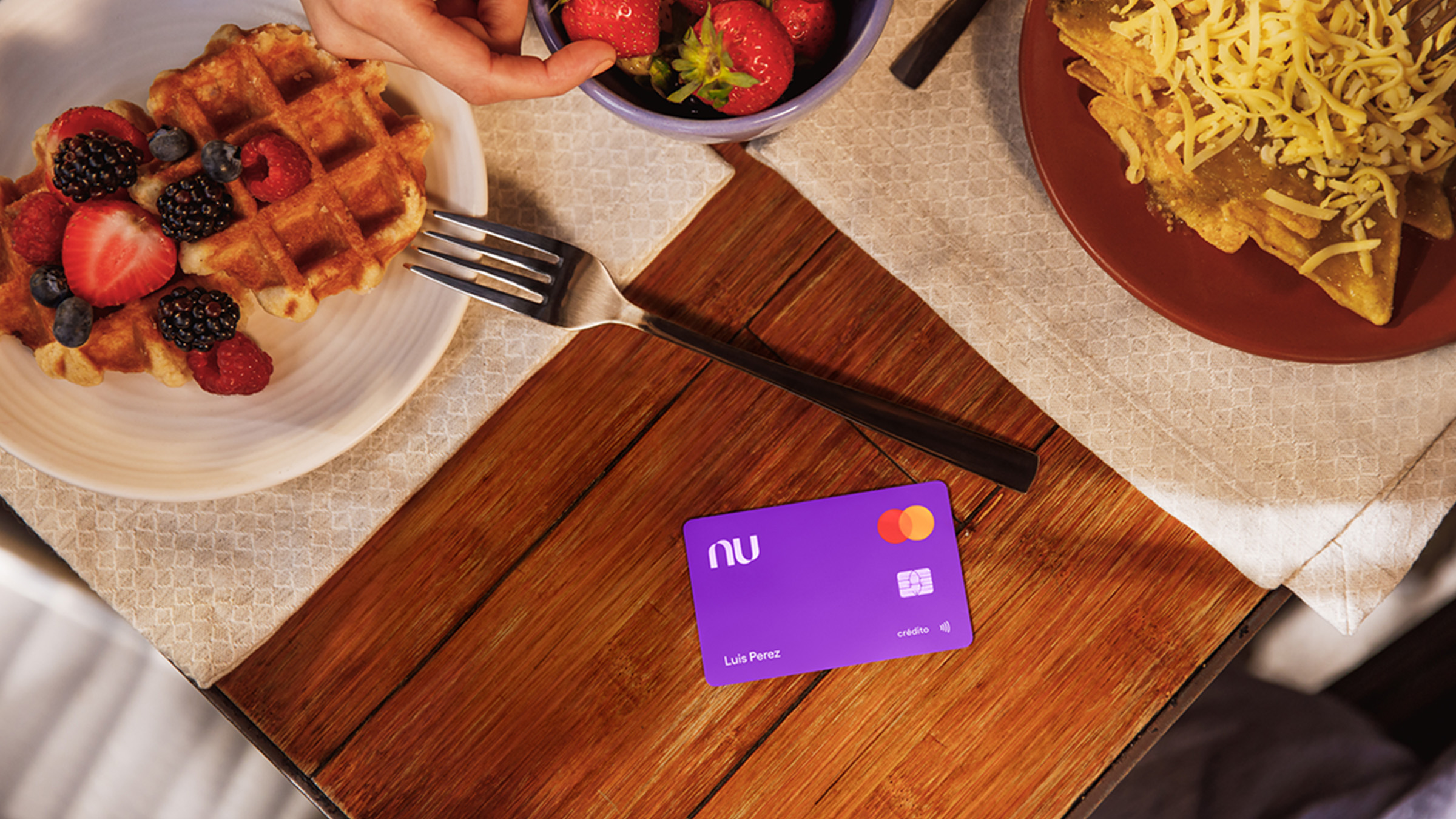 Una tarjeta color morado se encuentra en una mesa junto a platos de comida que tienen wafles y fresas e invitan a descubrir las Misiones de la Tarjeta Garantizada Nu