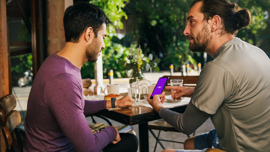 Dos hombres trigueños de cabello negro y ropa casual en tonos neutros están sentados platicando en la terraza de un restaurante y uno le explica al otro que fácil es solicitar una Tarjeta Nu en línea