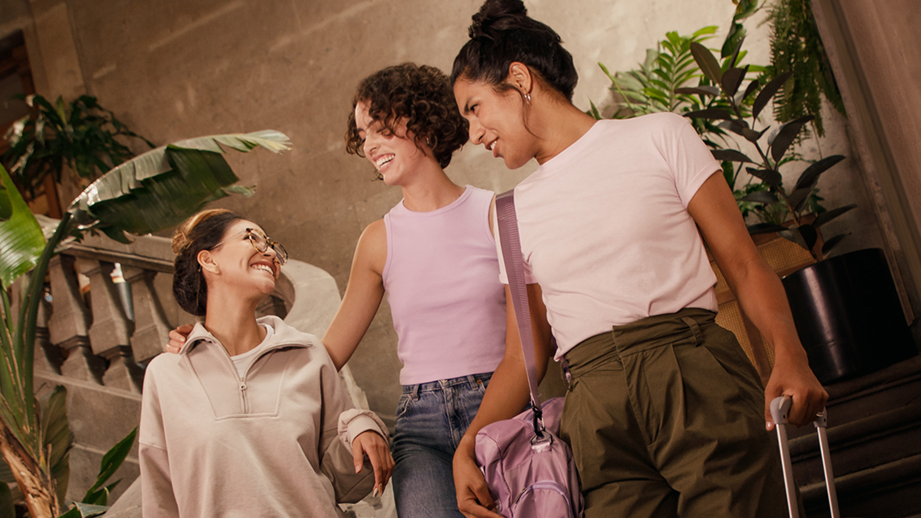Tres mujeres de edad mediana vestidas con ropas de colores neutros discuten acerca del crédito universitario en un patio interior que alberga algunas macetas con plantas.