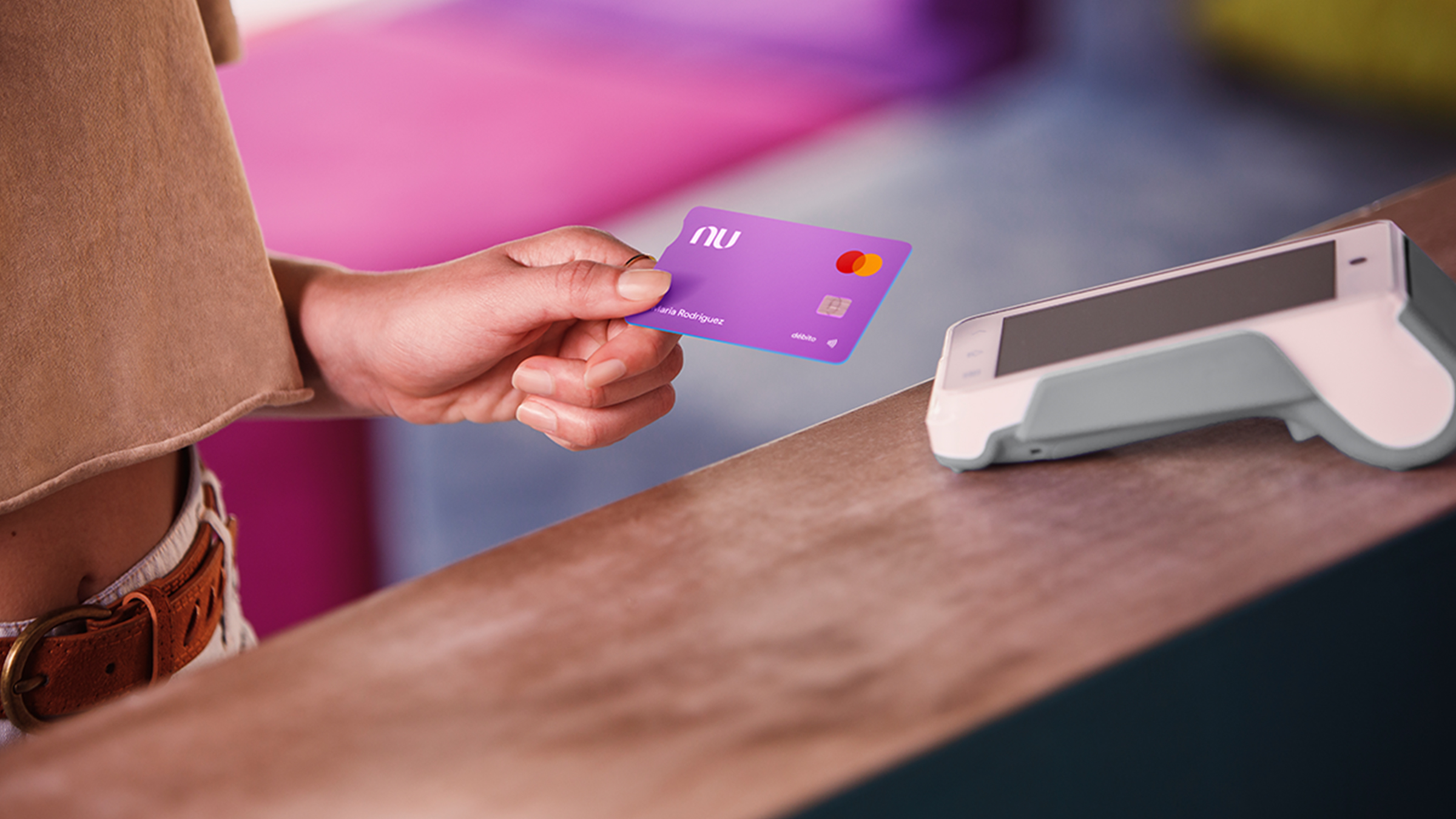 Una mano acerca la tarjeta de Cuenta Nu a una terminal punto de venta color blanca sobre un mostrador de madera en color nogal para hablara de las opiniones sobre cuenta nu