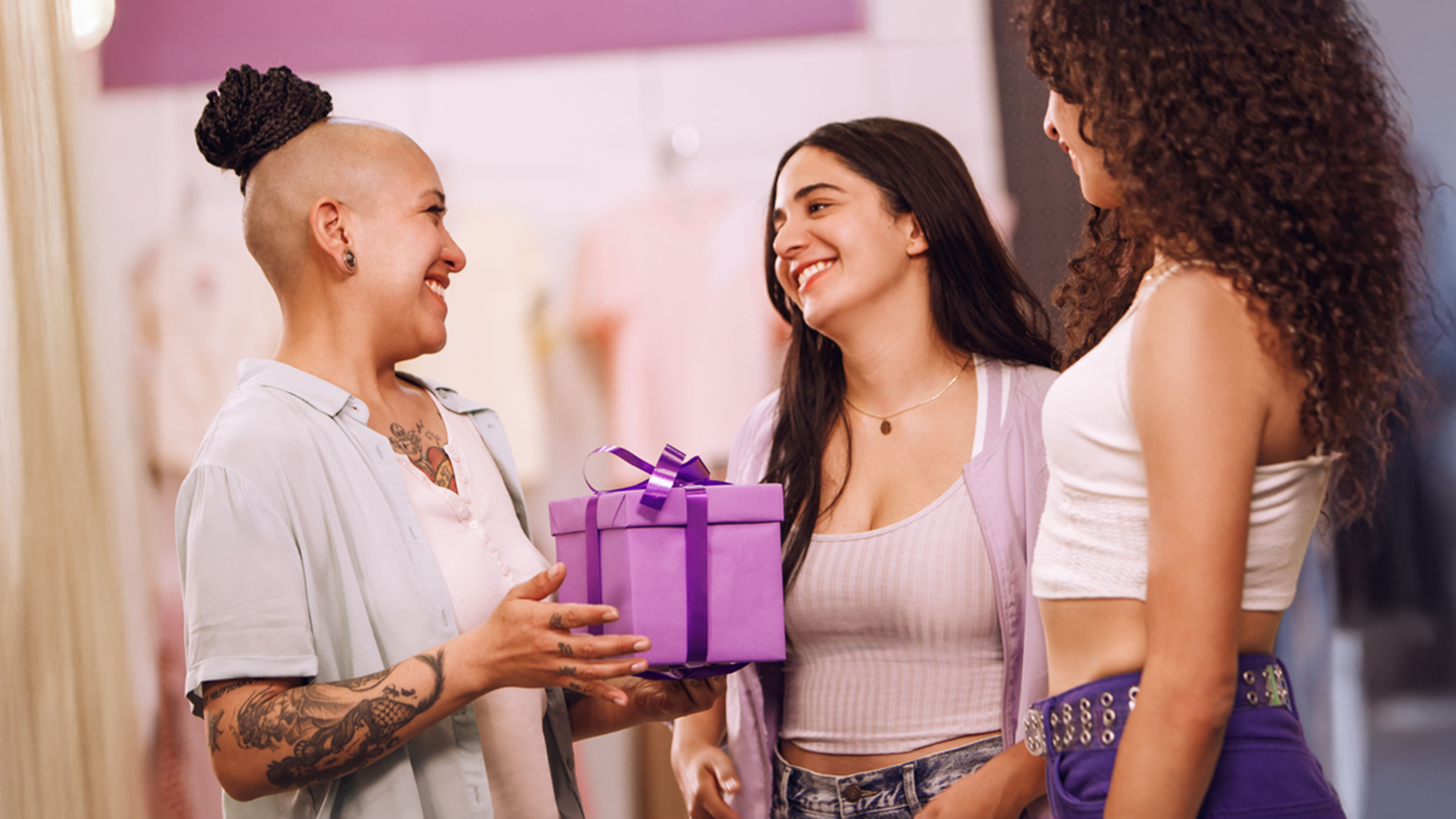 tres muejeres de origen latino vestidas con ropas claras y jeans dentro de una oficina sostien una caja de regalo color morado con un moño mientras comentan cómo sobrevivir al intercambio de regalos en la oficina