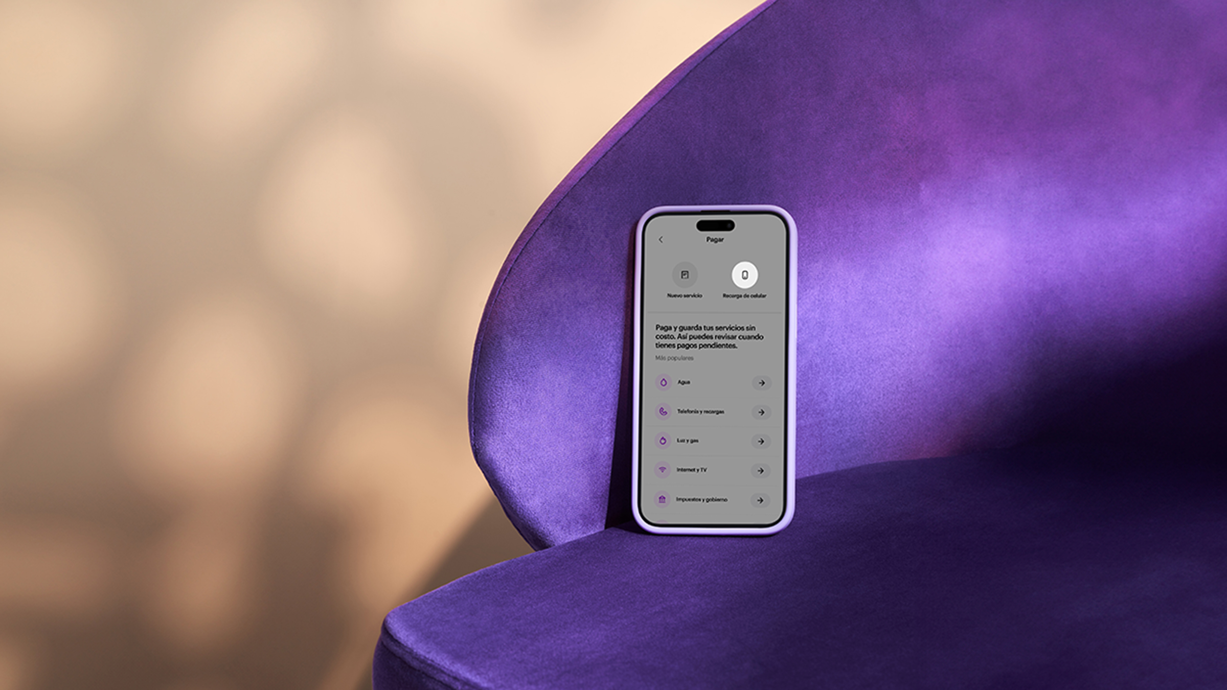 un teléfono móvil de color lila reposa sobre una silla de terciopelo morado en un ambiente iluminado por luz cálida para anunciar que ahora es posible recargar tu celular con Cuenta Nu