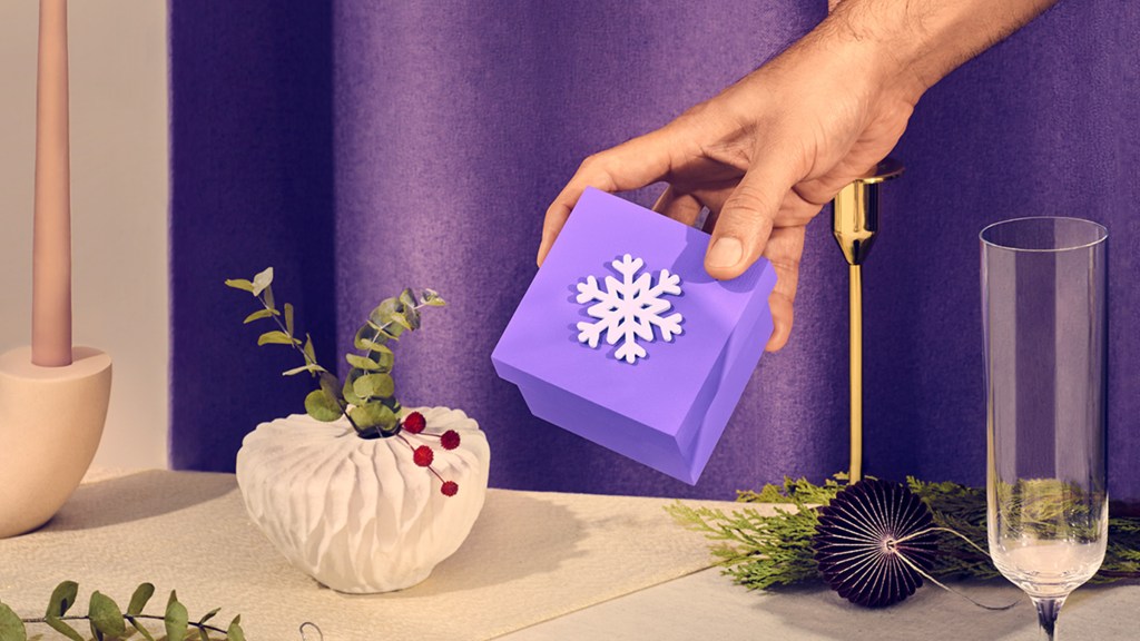 Una mano acerca una caja de regalo color morado decorada con un copo de nieve blanco en la tapa a una mesa decorada con un floraro con motivos navideños frente a una cortina morada que recuerda celebrar la navidad
