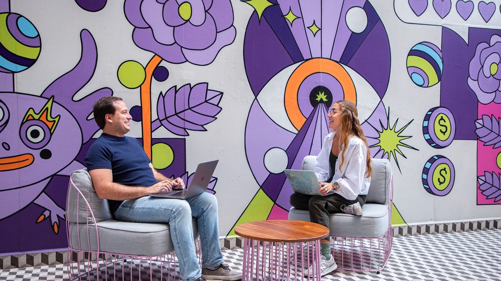 Una chica y un chico de mediana edad que visten jeans azul claro y ropa informal hablan sobre su carrera en Nu sentados en la sala de una terraza de las oficinas de Nu México adornadas con graffiti