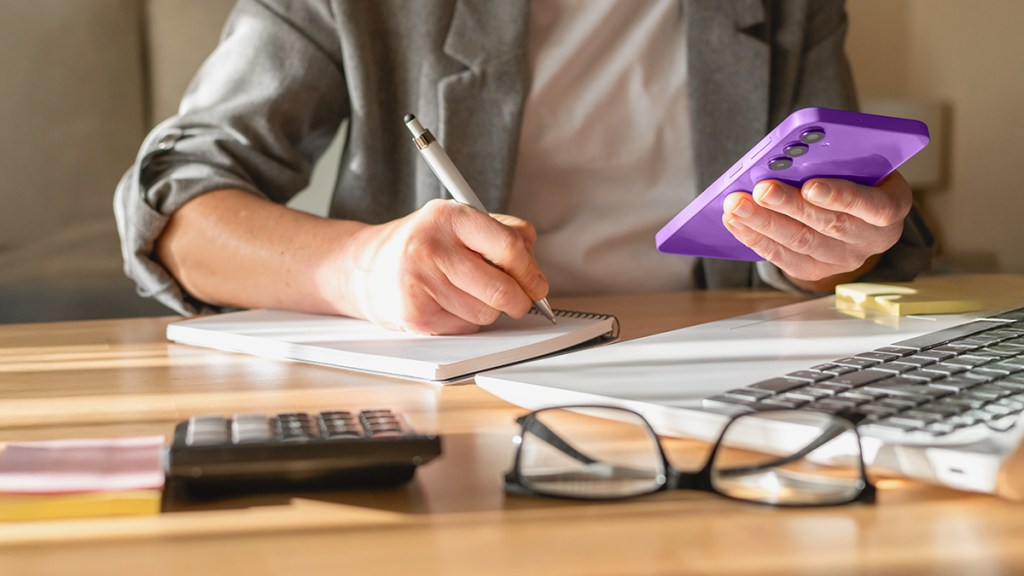 Persona tomando notas con calculadora y laptop en su escritorio, en el contexto de calcular si debe pagar impuestos por los rendimientos de Cuenta Nu.