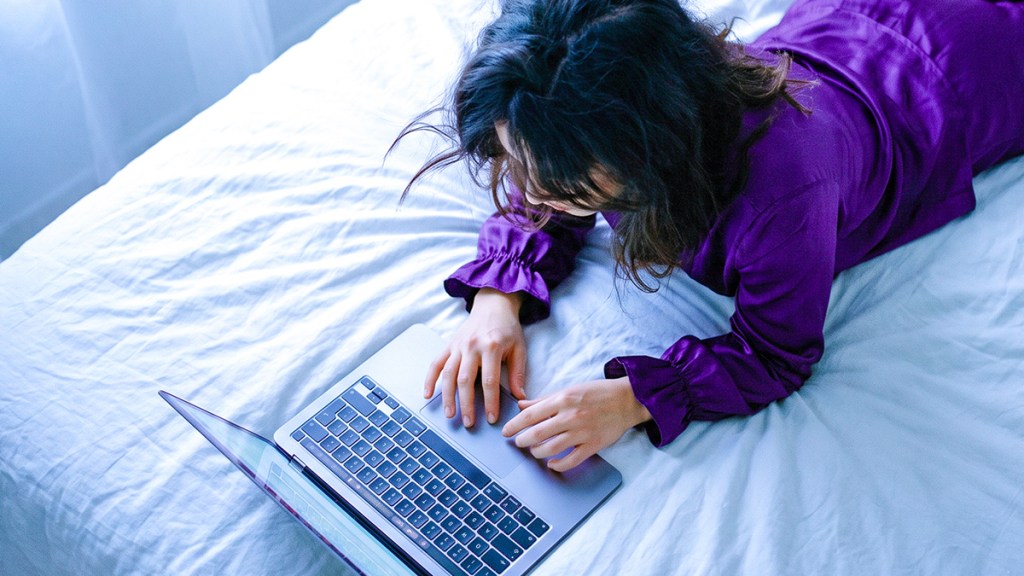 Persona usando su laptop en la cama con camisa morada, ilustrando cómo checar tu buró de crédito en línea de forma fácil para tomar control de tu historial crediticio.