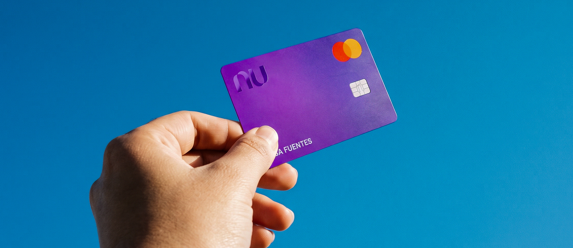 Mano sosteniendo su Tarjeta de Crédito Nu morada contra fondo azul, ilustrando cómo pagar una tarjeta de crédito completa antes de la fecha límite para evitar intereses.