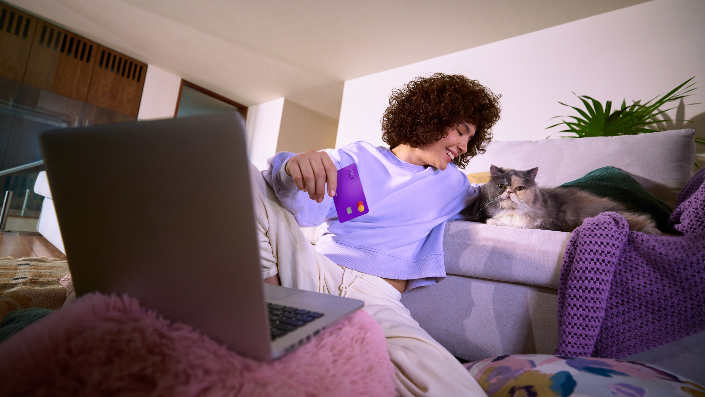Mujer cómoda en su sofá con su laptop y tarjeta morada, representando la tranquilidad de transferir saldo entre tus cuentas Nu.