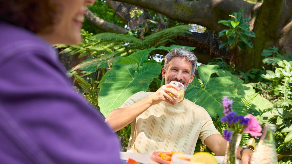 cajitas-nu-preguntas-respuestas_Header Hombre de cabello canoso sonriendo mientras bebe de un vaso, sentado en una mesa al aire libre rodeado de vegetación, con otra persona de blusa morada en primer plano.