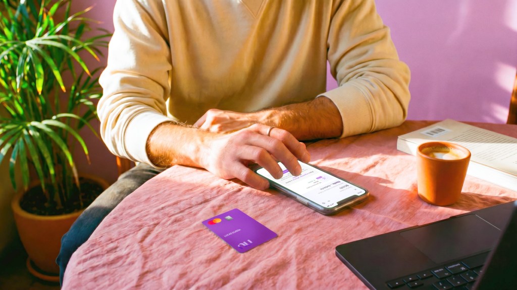 Persona revisando su app Nu en el celular con su tarjeta morada sobre el escritorio, ilustrando cómo decidir cuántas Cajitas Nu conviene tener para organizar tus ahorros.