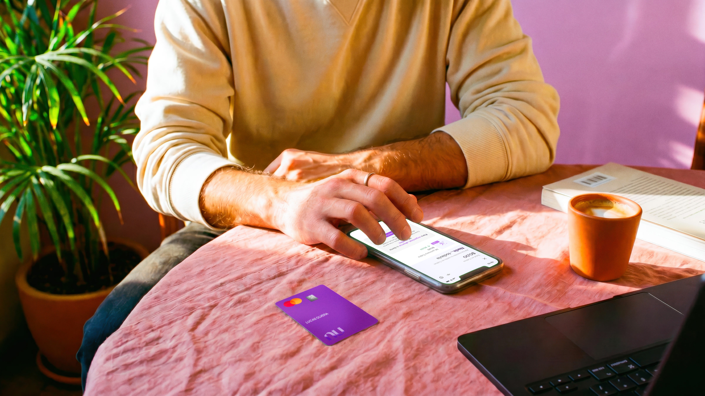 Persona revisando su app Nu en el celular con su tarjeta morada sobre el escritorio, ilustrando cómo decidir cuántas Cajitas Nu conviene tener para organizar tus ahorros.