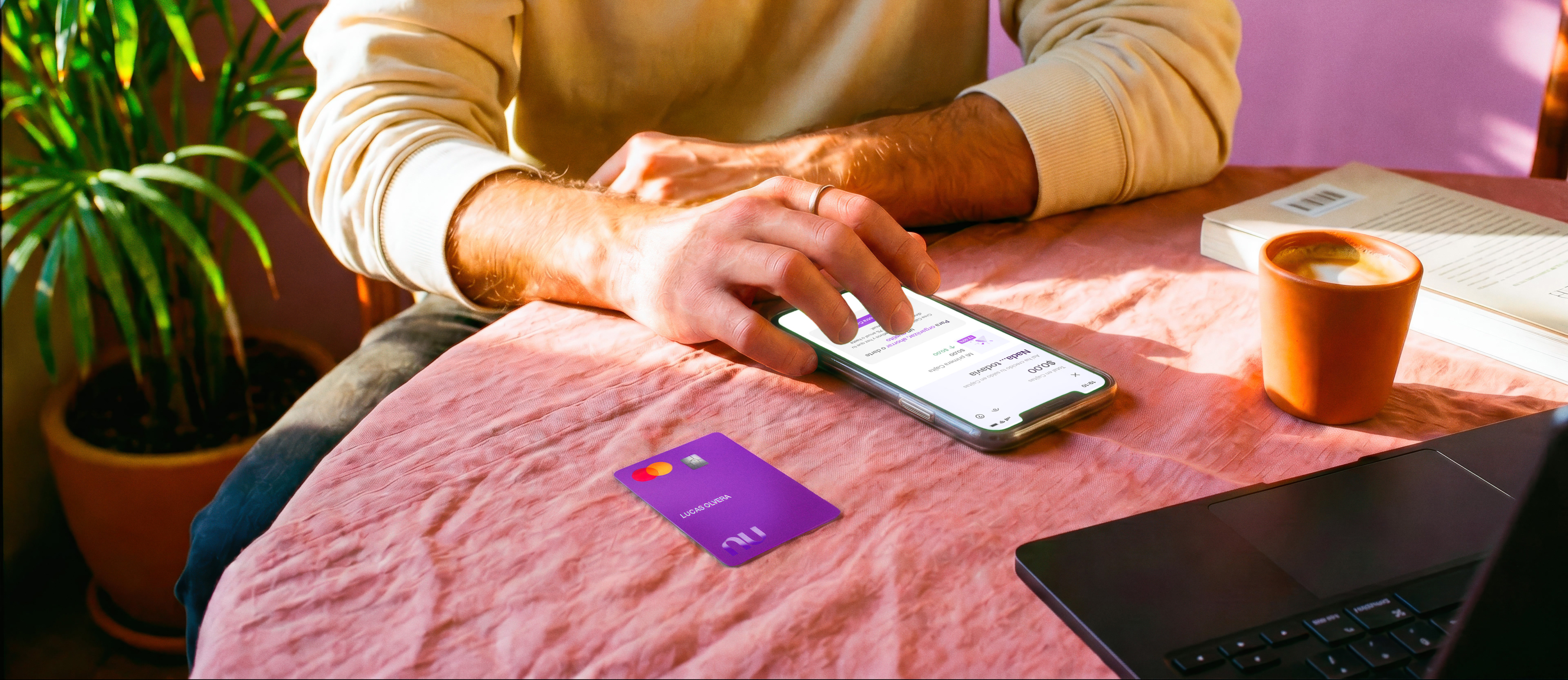 Persona revisando su app Nu en el celular con su tarjeta morada sobre el escritorio, ilustrando cómo decidir cuántas Cajitas Nu conviene tener para organizar tus ahorros.