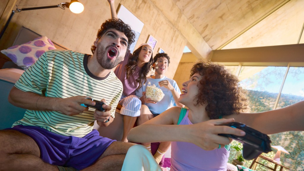 Grupo de amigos riendo y jugando videojuegos con controles en mano, en una sala con vista al bosque.