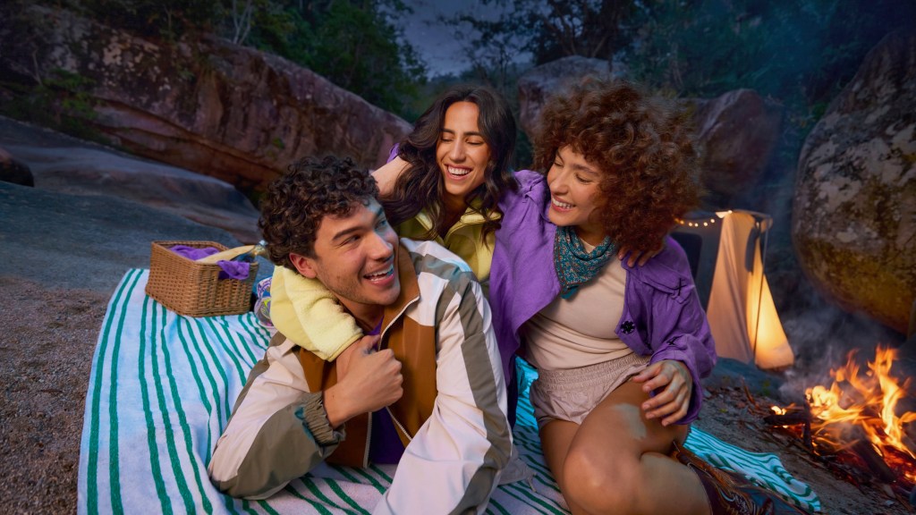 puedo-transferir-dinero-de-mi-tarjeta-de -credito_Header Tres amigos sonriendo y abrazados durante un picnic nocturno junto a una fogata, con una canasta y una manta de rayas de fondo.