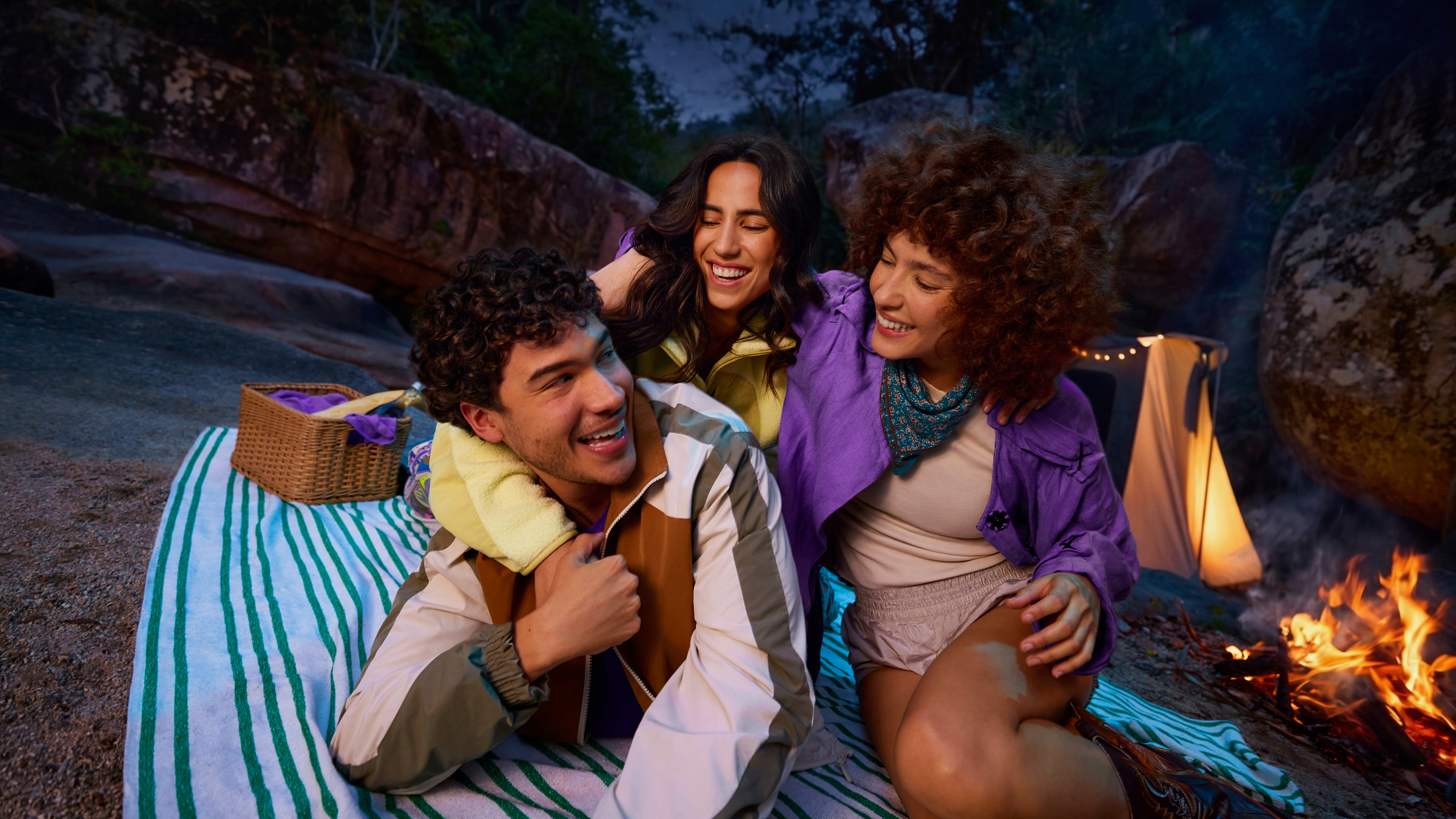 Tres amigos sonriendo y abrazados durante un picnic nocturno junto a una fogata, con una canasta y una manta de rayas de fondo.