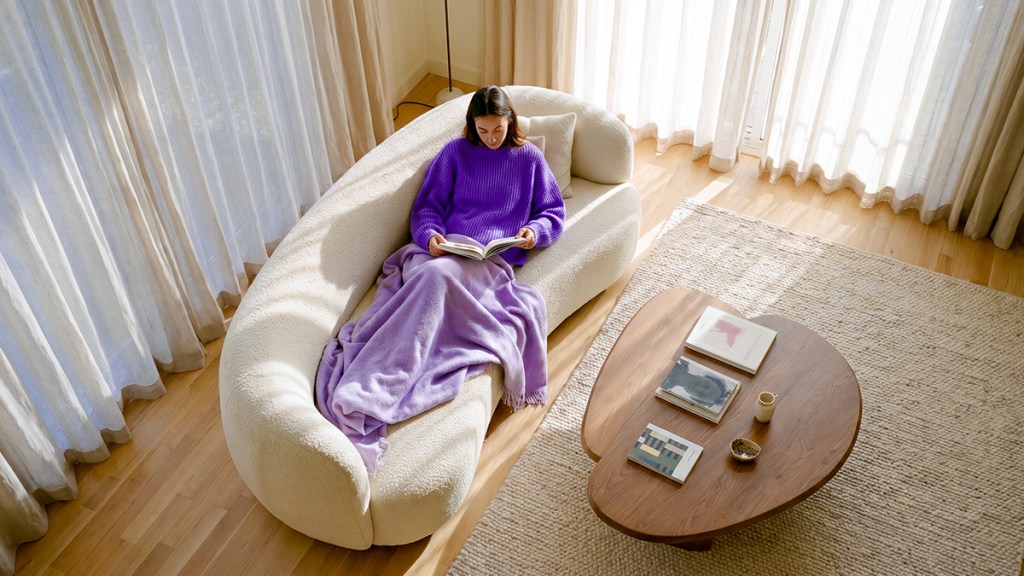 Mujer con suéter morado leyendo un libro en un sofá claro, cubierta con una cobija lila, en una sala con luz natural.