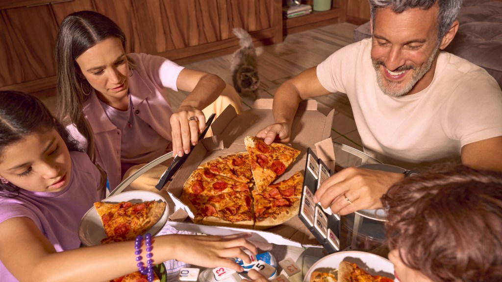 Familia disfrutando pizza en casa mientras juega un juego de mesa con fichas numeradas.