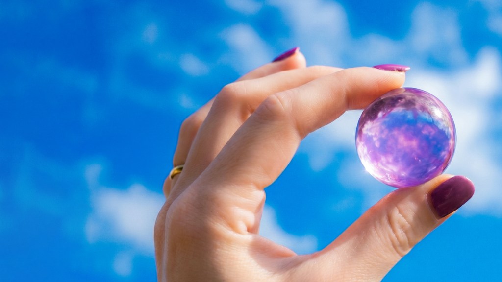 Persona sosteniendo una esfera morada translúcida contra un cielo azul claro con nubes blancas.