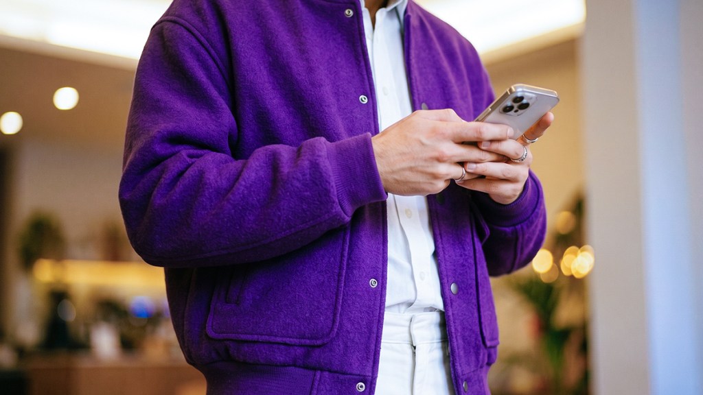 Persona con chamarra morada sosteniendo su celular, dentro de un espacio interior moderno con iluminación cálida.