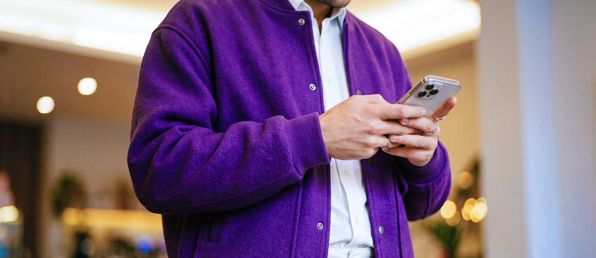 Persona con chamarra morada sosteniendo su celular, dentro de un espacio interior moderno con iluminación cálida.