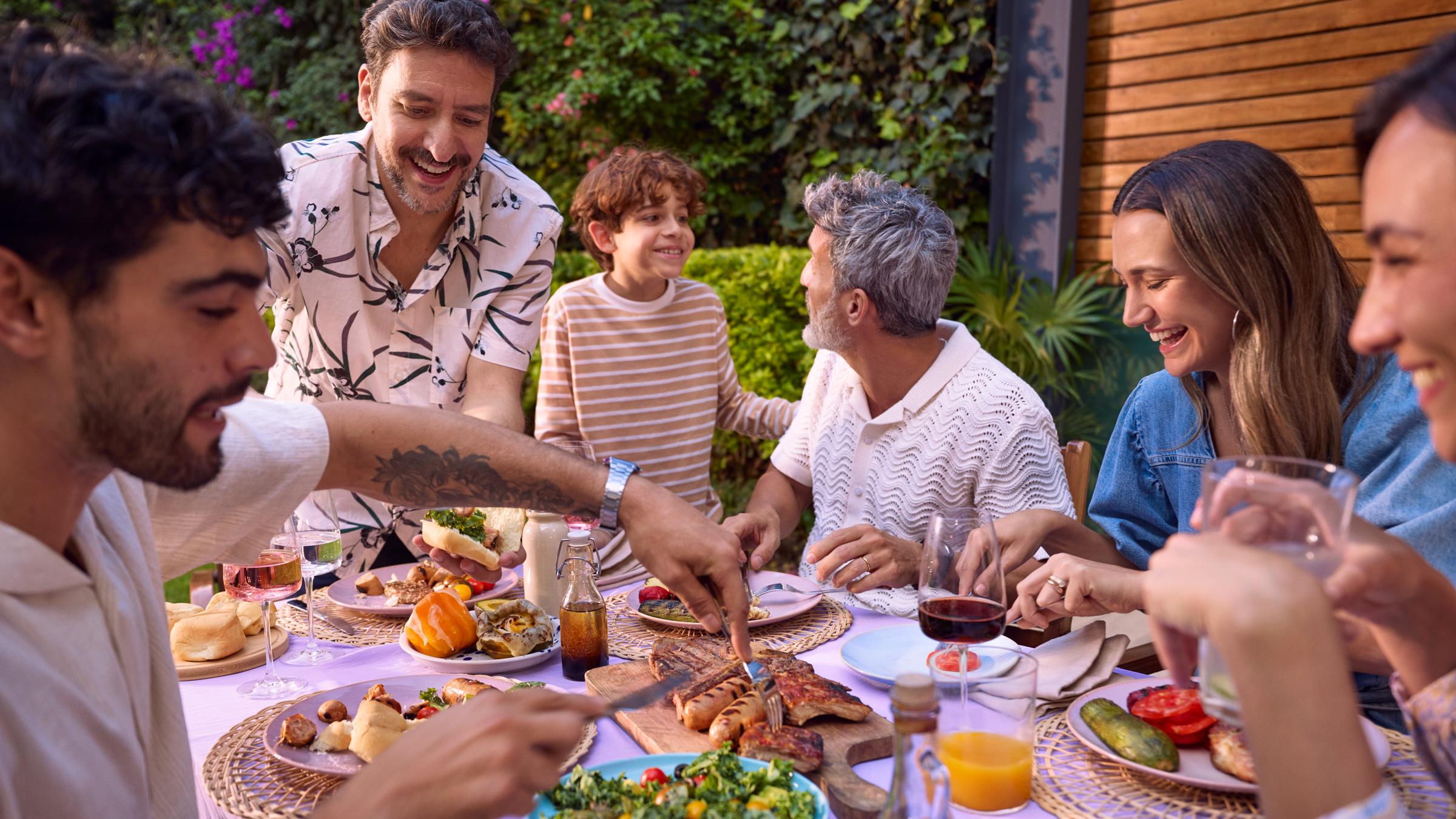 Familia multigeneracional disfrutando una comida al aire libre en un jardín, con comida, bebidas y conversación animada.