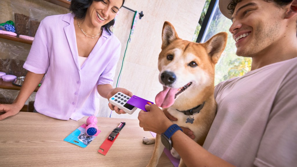 Dos personas sonriendo mientras realizan una transacción con Tarjeta de Crédito Nu en una terminal, con un perro corgi junto a ellos.