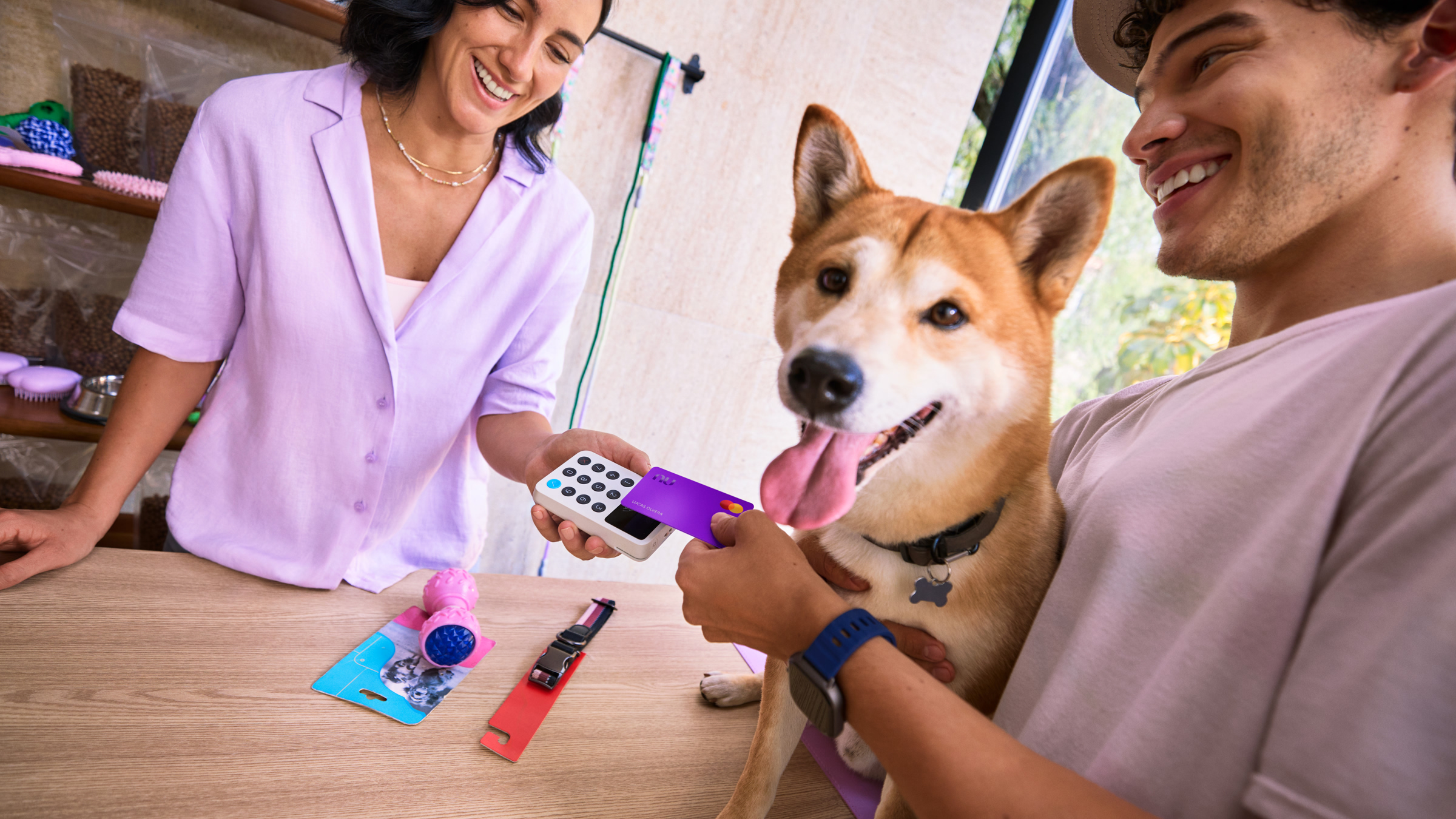 Dos personas sonriendo mientras realizan una transacción con Tarjeta de Crédito Nu en una terminal, con un perro corgi junto a ellos.