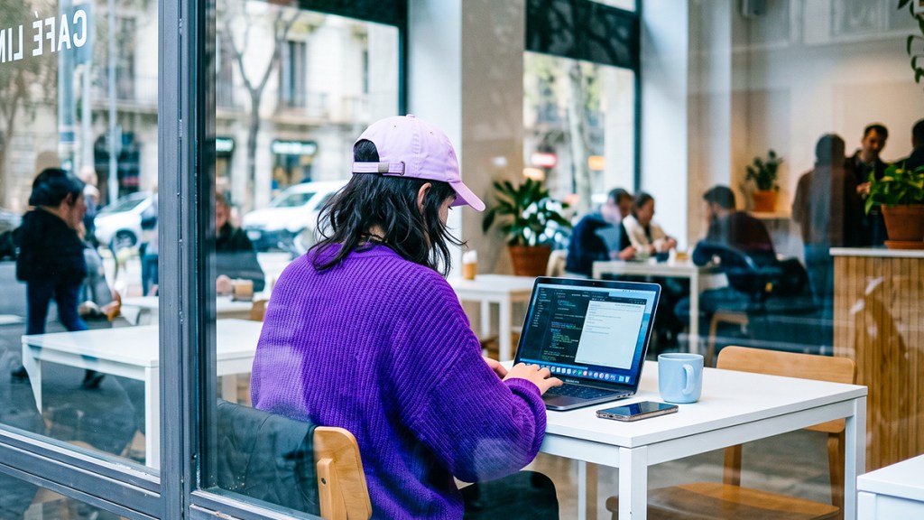 Persona con gorra morada trabajando en su laptop en una cafetería moderna, con un celular y taza sobre el escritorio.
