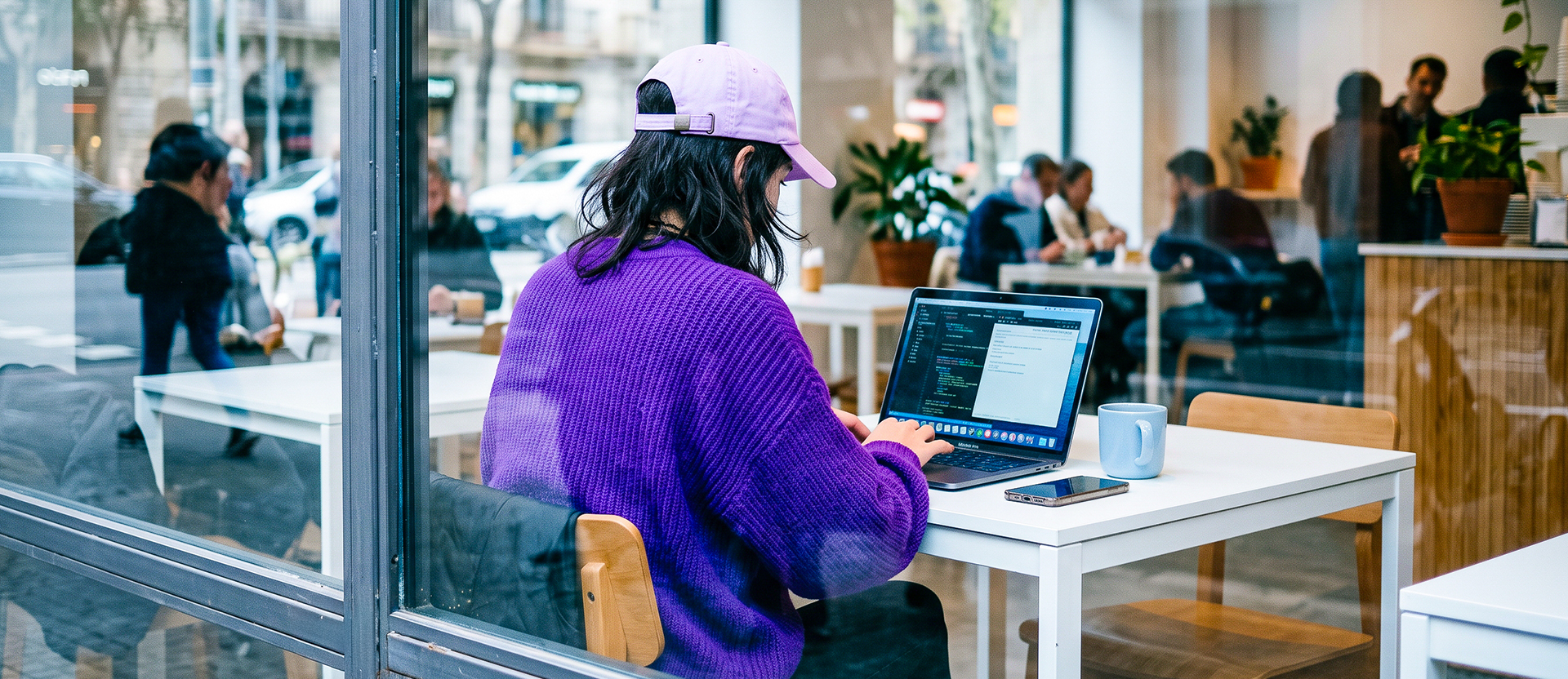 Persona con gorra morada trabajando en su laptop en una cafetería moderna, con un celular y taza sobre el escritorio.