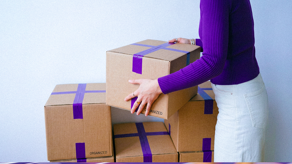 Persona organizando cajas etiquetadas "ORGANIZED" divididas con cintas moradas, ilustrando cómo administrar tu dinero con el método de los 3 apartados: gastos, ahorro y metas organizadas.