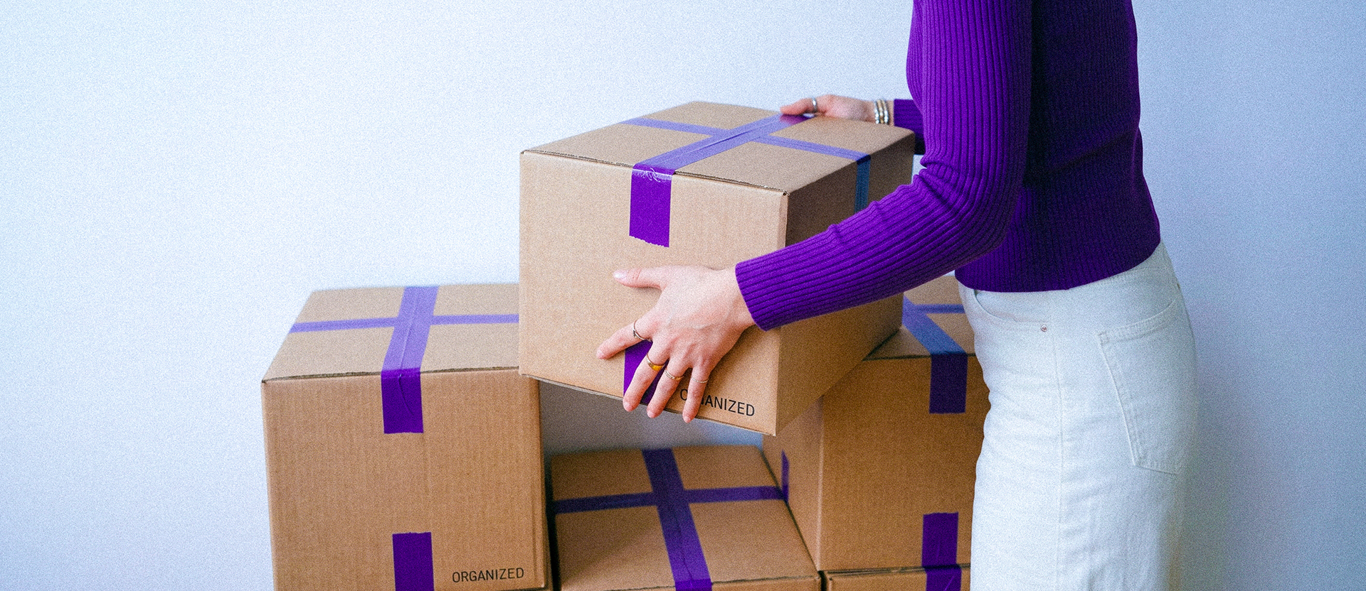 Persona organizando cajas etiquetadas "ORGANIZED" divididas con cintas moradas, ilustrando cómo administrar tu dinero con el método de los 3 apartados: gastos, ahorro y metas organizadas.