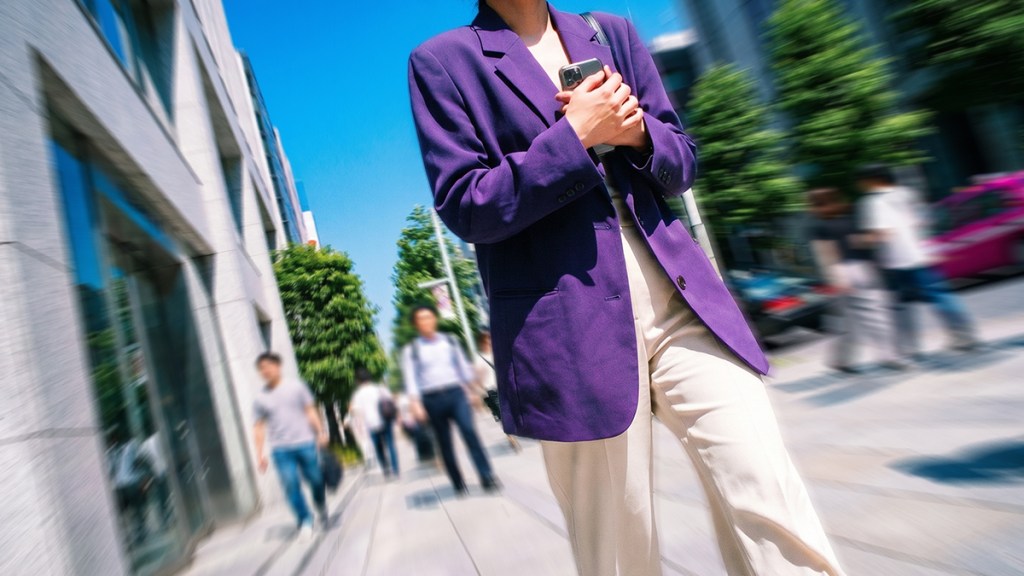 Persona con blazer morado sosteniendo su celular en una calle urbana, rodeada de gente y edificios.