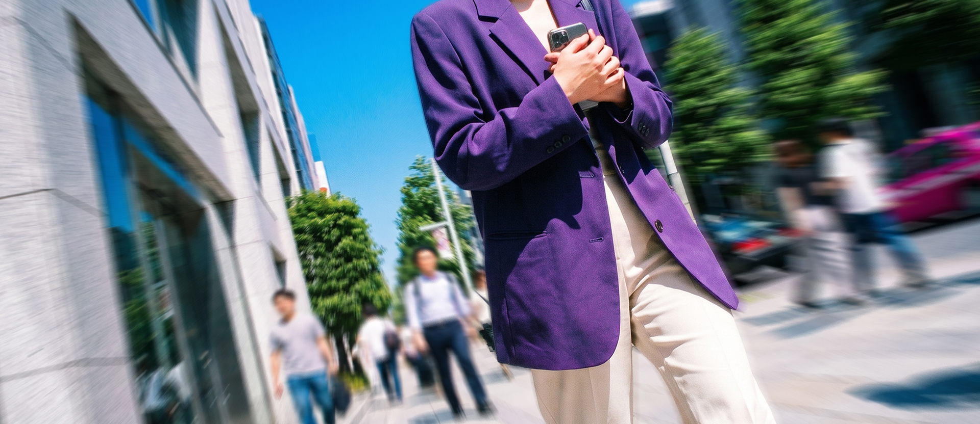 Persona con blazer morado sosteniendo su celular en una calle urbana, rodeada de gente y edificios.