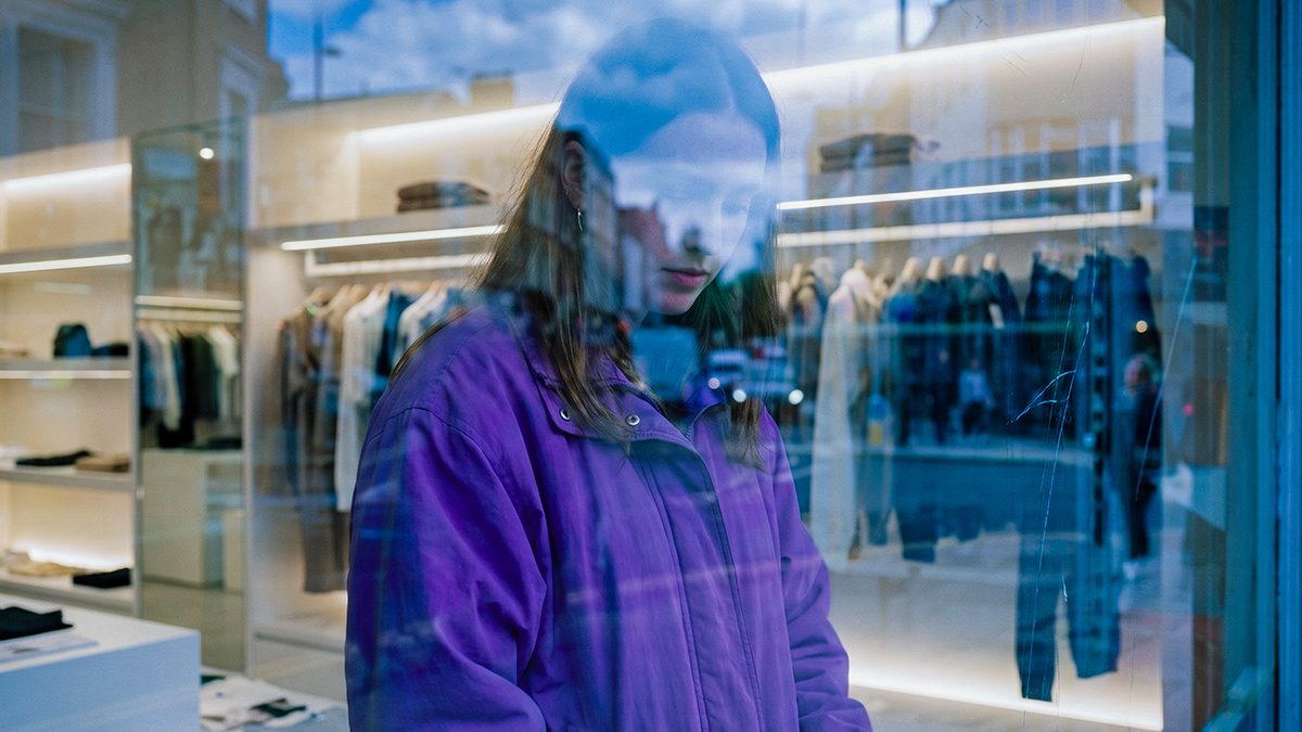 Mujer con chamarra morada frente al aparador de una tienda de ropa, pensativa antes de tomar una de sus decisiones financieras.