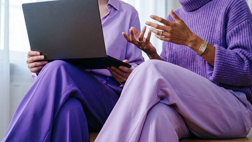 Dos personas con ropa morada conversan sentadas, una con laptop, sobre cómo denunciar fraudes por internet.