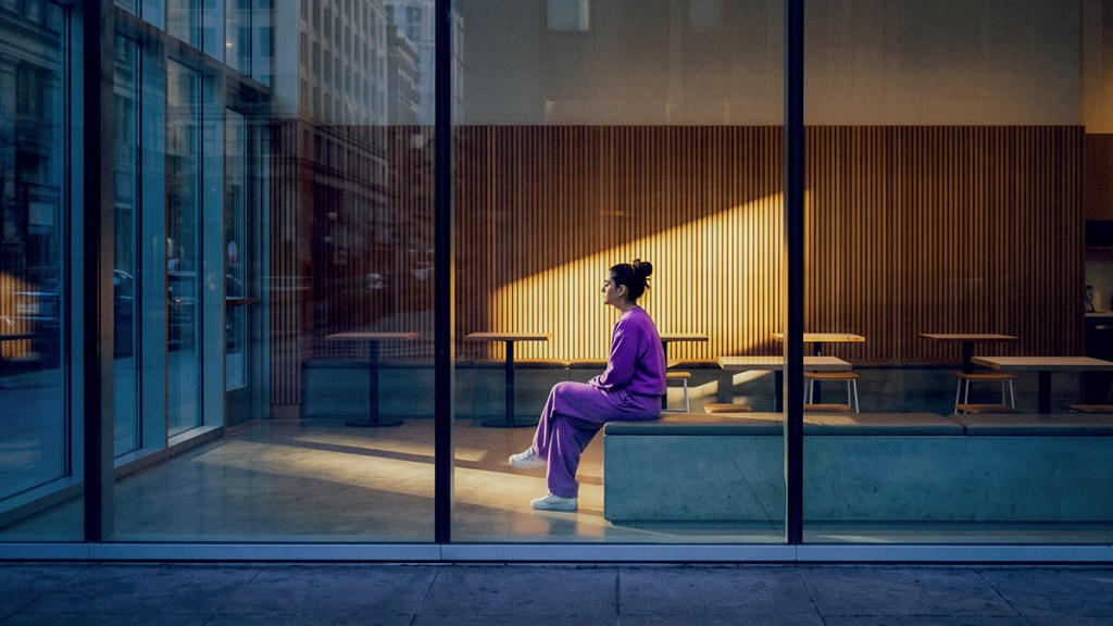 Persona con suéter morado sentada en un espacio moderno con ventanas de cristal, vista a la ciudad y luz natural dorada.